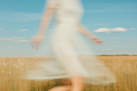 A blurred portrait of a young woman in a flowing white dress, joyfully running through golden grass under a bright blue sky, symbolizing freedom and vitalityの素材