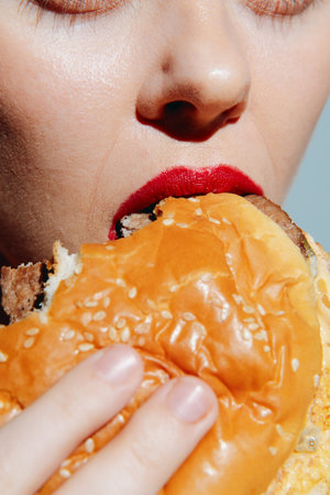 Close-up of a womans face taking a big bite of a juicy burger with red lipstick on, showcasing indulgence and craving for fast food.の写真素材