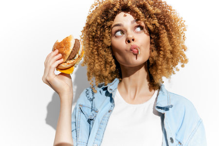 Woman with curly hair enjoying a burger, making a playful expression, showcasing a vibrant lifestyle and enticing fast food conceptの写真素材