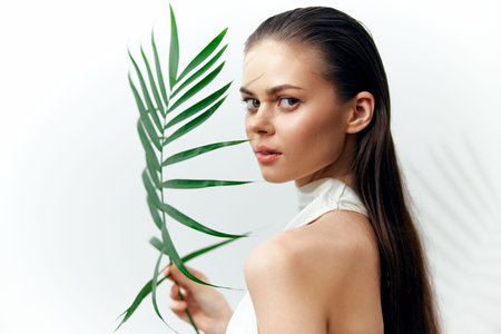 Young woman holding a green palm leaf, showcasing natural beauty with serene expression and minimalistic style against a white background.の写真素材