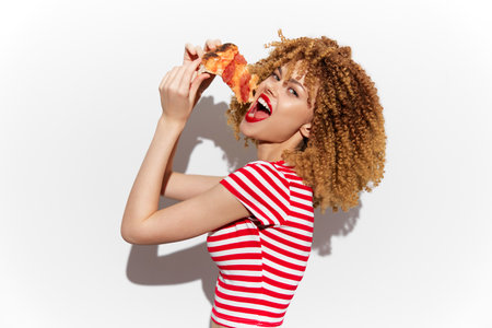 Young mixed race woman with curly hair enjoying pizza, colorful striped shirt, vibrant expression against a white backgroundの写真素材