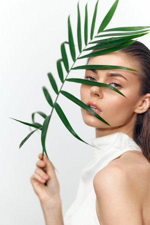A portrait of a young woman with natural beauty, holding a green palm leaf, conveying freshness and elegance in a minimalist setting.の写真素材