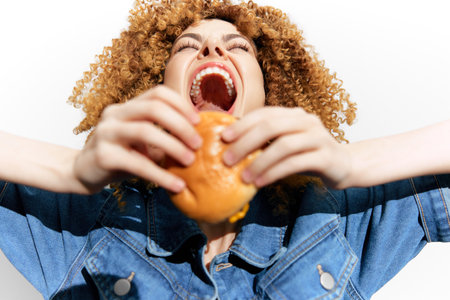 Happy woman with curly hair enjoying a delicious burger, wearing a denim jacket, joyful expression, bright background showcases the excitement of eatingの写真素材