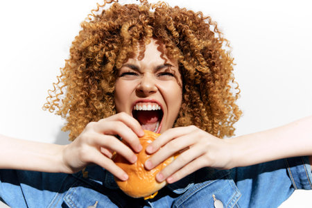 Young woman with curly hair joyfully biting into a burger, wearing denim, against a bright white background, conveying excitement and enjoyment of fast foodの写真素材