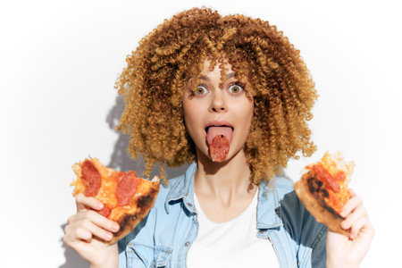 Playful young woman with curly hair joyfully eating pizza, showcasing a fun expression and vibrant personality, perfect for food and lifestyle themesの写真素材
