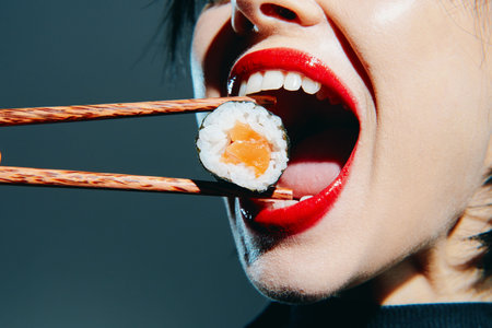 Close-up of a woman with red lips using chopsticks to lift sushi, emphasizing a cheerful and indulgent moment in modern Japanese cuisine.の写真素材