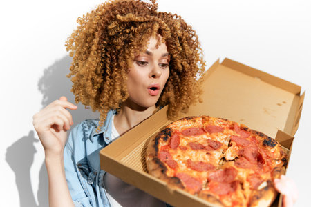 Excited young woman with curly hair looks at delicious pizza in takeaway box, expressing joy and surprise, perfect for food and lifestyle conceptsの写真素材