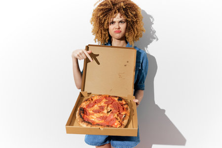 Displeased young woman with curly hair holding an unappetizing pizza in a box, showcasing her disappointment in the food qualityの写真素材