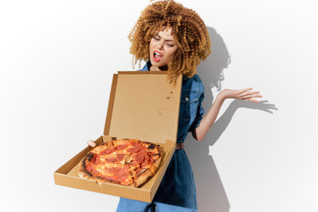 A young woman with curly hair holds a pizza box, expressing surprise and delight, wearing a denim outfit against a clean white backgroundの写真素材
