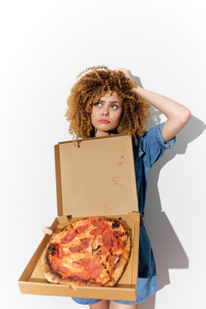 Young woman with curly hair holding a pizza box, looking thoughtful Expression conveys a mix of desire and concern about eating pizzaの写真素材