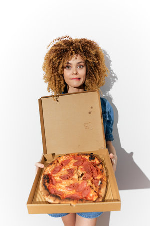 Young woman with curly hair joyfully holding a pizza box, showcasing vibrant pizza, against a minimalist white background, perfect for food related themesの写真素材