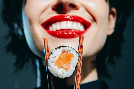 Smiling woman holding sushi with red lips against a dark background, showcasing Japanese cuisine....