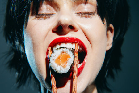 A young woman with short black hair passionately eating sushi using chopsticks, showcasing excitement and joy in her expression against a dark backdrop.の写真素材