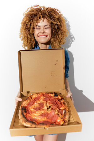 Young woman with curly hair happily holding a pizza box, showcasing a delicious pepperoni pizza against a bright white background, perfect for food loversの写真素材