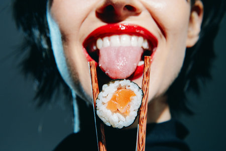 Playful female model with sushi on chopsticks, showcasing vibrant lips and tongue, in a modern, stylish setting.の写真素材