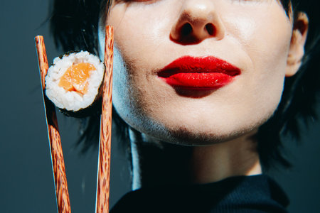 Stylish woman holding sushi with chopsticks, vibrant red lips, dark background, showcasing modern culinary delights and cultural fusion.の写真素材
