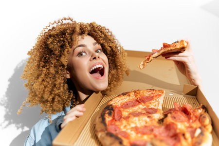 Young diverse woman joyfully holding a pizza box and slice with curly hair, showcasing excitement and deliciousness in a bright, minimalist settingの写真素材
