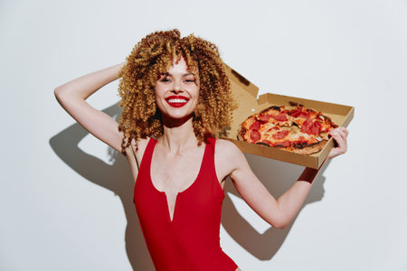 Joyful young woman holding a pizza box, wearing a red swimsuit, showcasing vibrant hair and bright smile against a plain white backgroundの写真素材