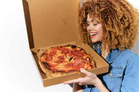 Smiling woman with curly hair holding a delicious pepperoni pizza in a takeout box, showcasing joy and excitement in a bright settingの写真素材