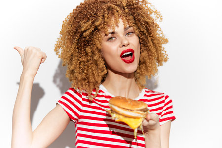 Cheerful young woman with curly hair displaying a delicious cheeseburger, wearing a red and white striped shirt, perfect for showcasing tasty fast foodの写真素材