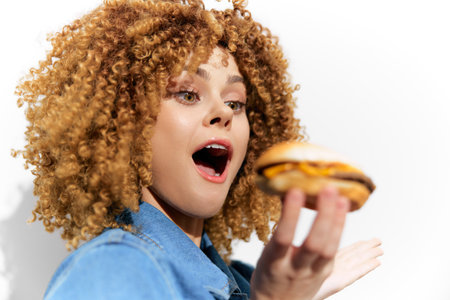 Excited young woman with curly hair holding a delicious cheeseburger, expressing joy and enthusiasmの写真素材