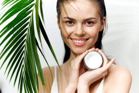 Smiling model holding skincare cream with tropical greenery background, showcasing beauty and freshness in wellness concept.の写真素材