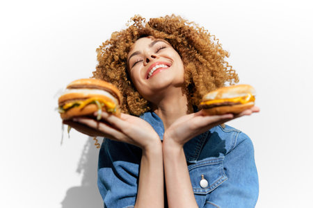 Happy young woman with curly hair joyfully holding two delicious burgers, showcasing vibrant colors and a fun food concept in a bright backgroundの写真素材