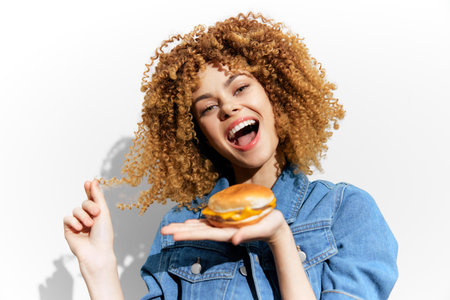 Happy young woman with curly hair holding a cheeseburger, smiling joyfully against a bright white background, showcasing a fun and casual vibeの写真素材