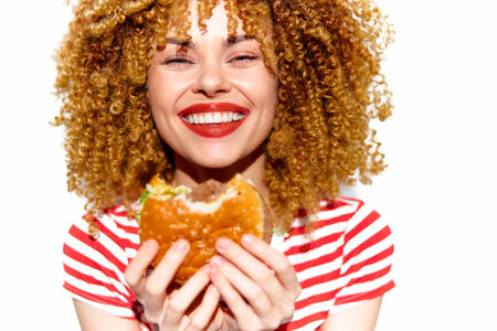 Young woman with curly hair smiling while holding a delicious burger, wearing a striped shirt, representing a joyful moment in fast food enjoymentの写真素材