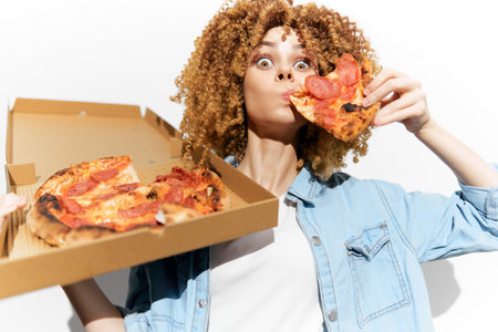 Young woman with curly hair excitedly eating pizza, showcasing a fun and joyful moment, emphasizing delicious food and casual style, perfect for food related conceptsの写真素材
