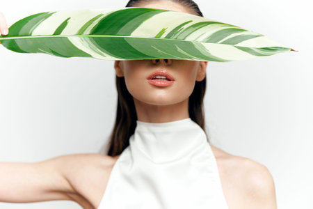 Elegant model concealing her face with a large green leaf, showcasing a serene expression and minimalist style in a studio setting.の写真素材