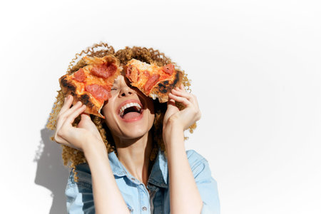 Joyful woman with curly hair playfully holding pizza slices against a white background, expressing...