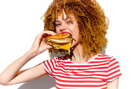Young woman with curly hair enjoying a delicious burger, wearing a striped red and white shirt, vibrant colors highlight a joyful eating conceptの写真素材