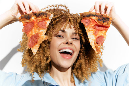 Happy young woman with curly hair holding slices of pepperoni pizza, celebrating with a joyful smile...