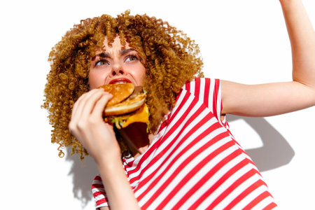 Young woman with curly hair joyfully eating a burger, wearing a striped shirt, against a bright white background, symbolizing happiness and indulgence in fast foodの写真素材