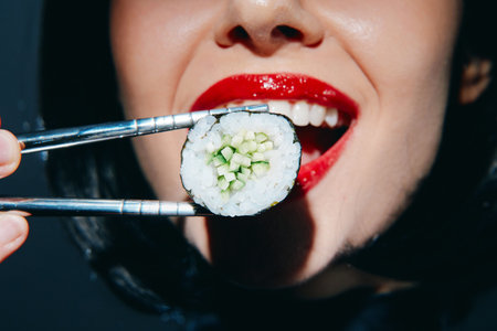 Close-up of a woman with red lipstick holding sushi with chopsticks, showcasing vibrant colors and a...