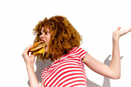 Young woman with curly hair in a striped shirt, joyfully eating a burger against a white background, embodying the concept of indulgent fast food enjoymentの写真素材