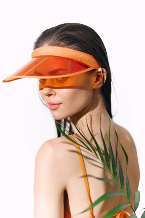 Young woman with an orange visor and tropical leaves, expressing confidence and style against a minimalistic background.の写真素材