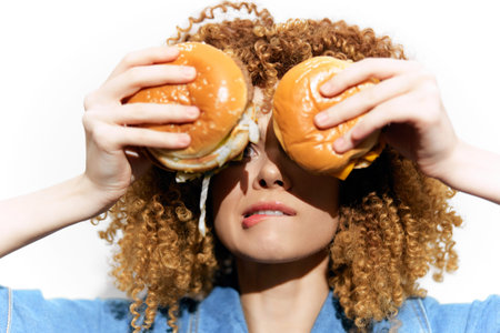 Young woman with curly hair playfully holding burgers in front of her face, showcasing a fun and...