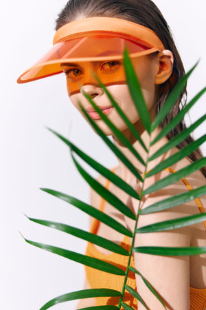Young woman in vibrant orange outfit with visor, hiding behind a green palm leaf, exuding summer vibes and playful emotion.の写真素材
