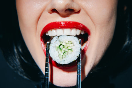 Close-up of a woman with red lipstick joyfully holding sushi with chopsticks, showcasing vibrant colors and an energetic atmosphere.の写真素材