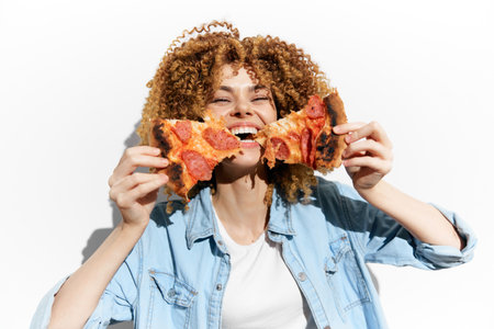 A joyful young woman with curly hair enjoying slices of pizza, showcasing her love for food in a light hearted, casual setting with a bright backgroundの写真素材
