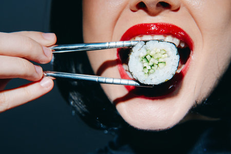 A close-up shot of a womans mouth with bold red lips joyfully holding sushi with chopsticks,...