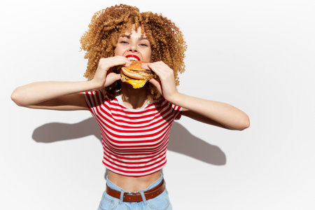 Happy young woman with curly hair enjoying a delicious hamburger, wearing a striped shirt and jeans against a white background, showcasing vibrant energy and joyの写真素材