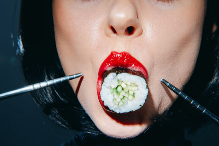 Close-up of a woman with red lips savoring sushi, using chopsticks, against a dark background highlighting a modern and bold culinary concept.の写真素材