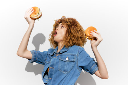 Excited young woman holding burgers, celebrating food with joyful expression and stylish denim outfit against a minimalist backgroundの写真素材