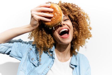 Cheerful young woman with curly hair enjoying a burger, bright and colorful background showcasing food happiness and a casual styleの写真素材