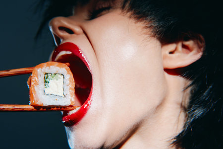 Woman with red lips enjoying sushi, showcasing vibrant emotions and modern lifestyle against a dark background. A fusion of taste and expression.の写真素材
