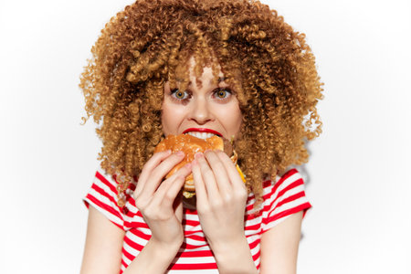 Cheerful young woman with curly hair enjoying a delicious burger, wearing a red and white striped shirt, brightening the scene with her vibrant energy and playful emotionsの写真素材