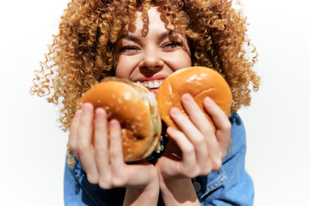 Happy young woman with curly hair joyfully holding two burgers against a bright white background, celebrating the delight of eating and indulgenceの写真素材